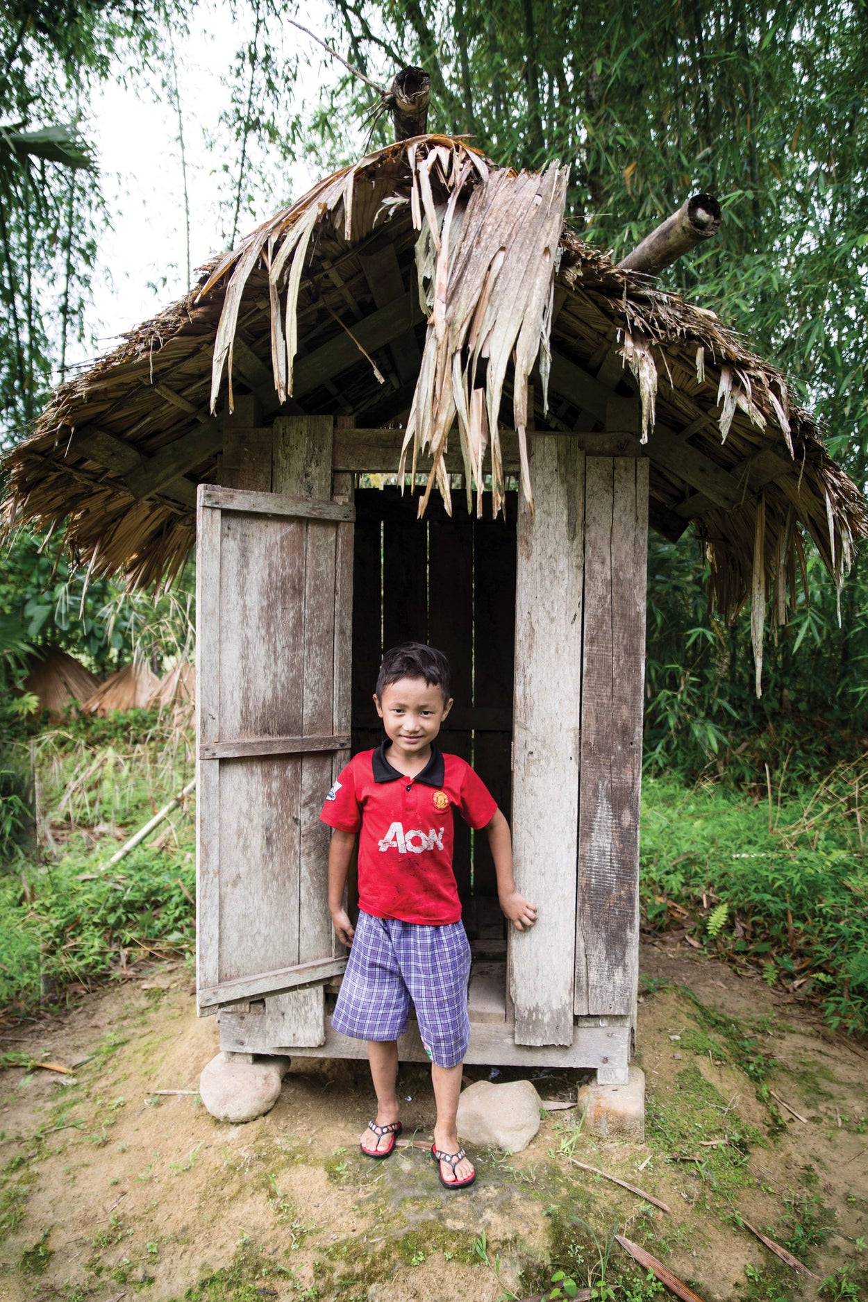 boy in a red shirt standing in front of a latrine